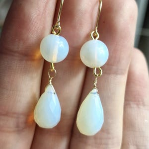 May include: A pair of gold dangle earrings with white opal stones. The earrings feature a round opal bead at the top and a teardrop-shaped opal bead at the bottom.