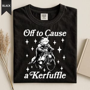Könnte beinhalten: Schwarzes T-Shirt mit einem weißen Aufdruck einer Froschfigur auf einem Fahrrad, mit Umhang und Sternen. Der Text lautet "Off to Cause a Kerfuffle". Ein lässiges, neuartiges Design.