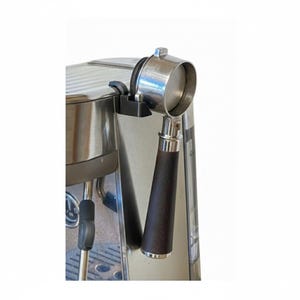 以下が含まれることがあります： ステンレス鋼のエスプレッソマシン。ポルタフィルターが取り付けられています。ポルタフィルターは濃い茶色の木製ハンドルと銀色の金属製フィルターバスケットを備えています。マシンはシルバー仕上げで、黒いプラスチック部品が付いています。