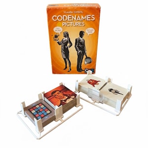 Op de afbeelding: Bordspel "Codenames Pictures" met een oranje doos met illustraties van een man en een vrouw. Het spel bevat kaartspellen in witte plastic houders, met verschillende afbeeldingen op de kaarten.