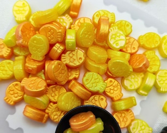 Può includere: Una collezione di piccoli ciondoli a forma di pesce nelle tonalità del giallo e dell'arancione con una finitura metallica perlata. Alcuni ciondoli sono sparsi su uno stampo in silicone bianco con bordi smerlati, altri in un piccolo misurino nero.