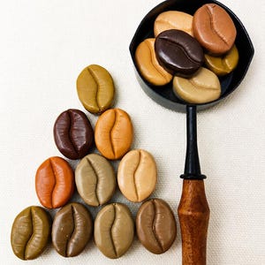 May include: A collection of smooth, oval-shaped coffee bean replicas in various shades of brown, tan, and beige. Several beans are arranged in a triangular formation, while others are contained within a small black metal scoop with a wooden handle.