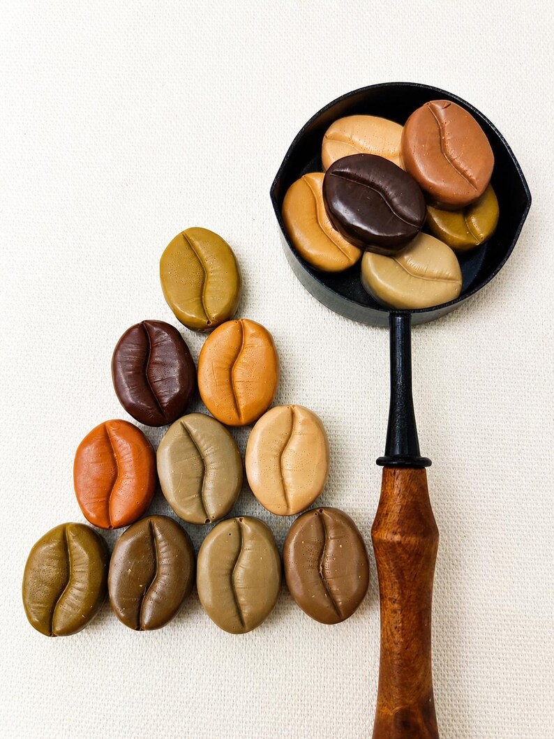 May include: A collection of smooth, oval-shaped coffee bean replicas in various shades of brown, tan, and beige. Several beans are arranged in a triangular formation, while others are contained within a small black metal scoop with a wooden handle.