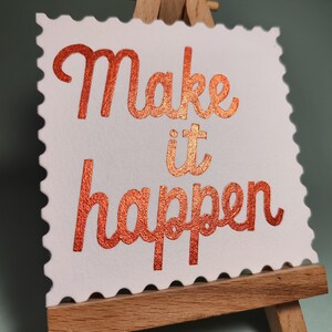 Pu&ograve; includere: Una carta bianca con bordo smerlato, che mostra le parole "Make it happen" in una scritta color rame e glitterata. La carta &egrave; appoggiata su un piccolo cavalletto di legno, ideale per l'arredamento della scrivania.