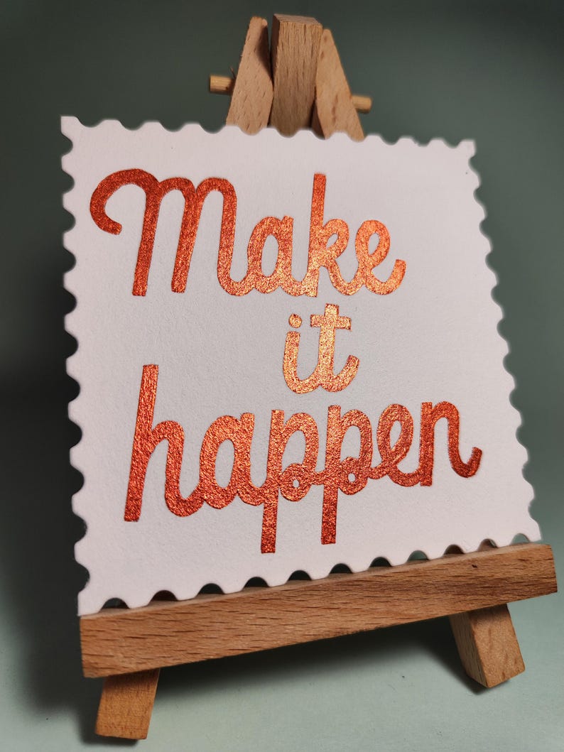 Pu&ograve; includere: Una carta bianca con bordo smerlato, che mostra le parole "Make it happen" in una scritta color rame e glitterata. La carta &egrave; appoggiata su un piccolo cavalletto di legno, ideale per l'arredamento della scrivania.