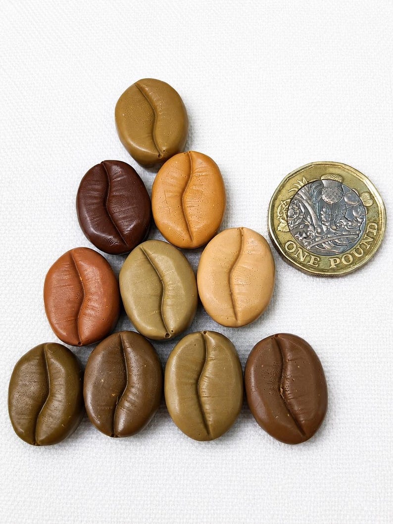 May include: A collection of nine oval-shaped, coffee bean-inspired objects in various shades of brown, ranging from light tan to dark chocolate. A British one-pound coin is positioned to the right for scale, showing its metallic texture and detail.