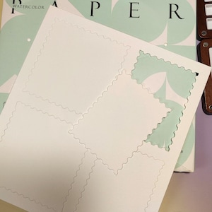 Puede incluir: Una pila de papel de acuarela blanco con un diseño perforado, listo para ser rasgado en hojas individuales. El papel está apilado encima de un bloc de papel de acuarela con la palabra "WATERCOLOR" visible. El fondo presenta un diseño verde claro y blanco.