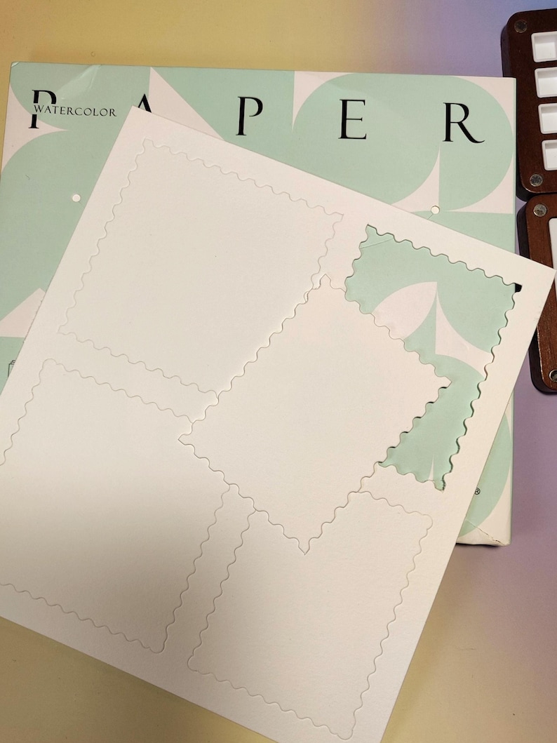 Pu&ograve; includere: Una pila di carta per acquerello bianca con un design perforato, pronta per essere strappata in fogli singoli. La carta &egrave; impilata sopra un blocco di carta per acquerello con la parola "WATERCOLOR" visibile. Lo sfondo presenta un design verde chiaro e bianco.