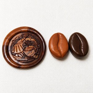 May include: A circular brown wax seal stamp featuring an embossed design of a coffee pot, a cup, and spices, accompanied by two smooth, oval-shaped coffee bean replicas, one in a light tan colour and the other in a dark chocolate brown colour.