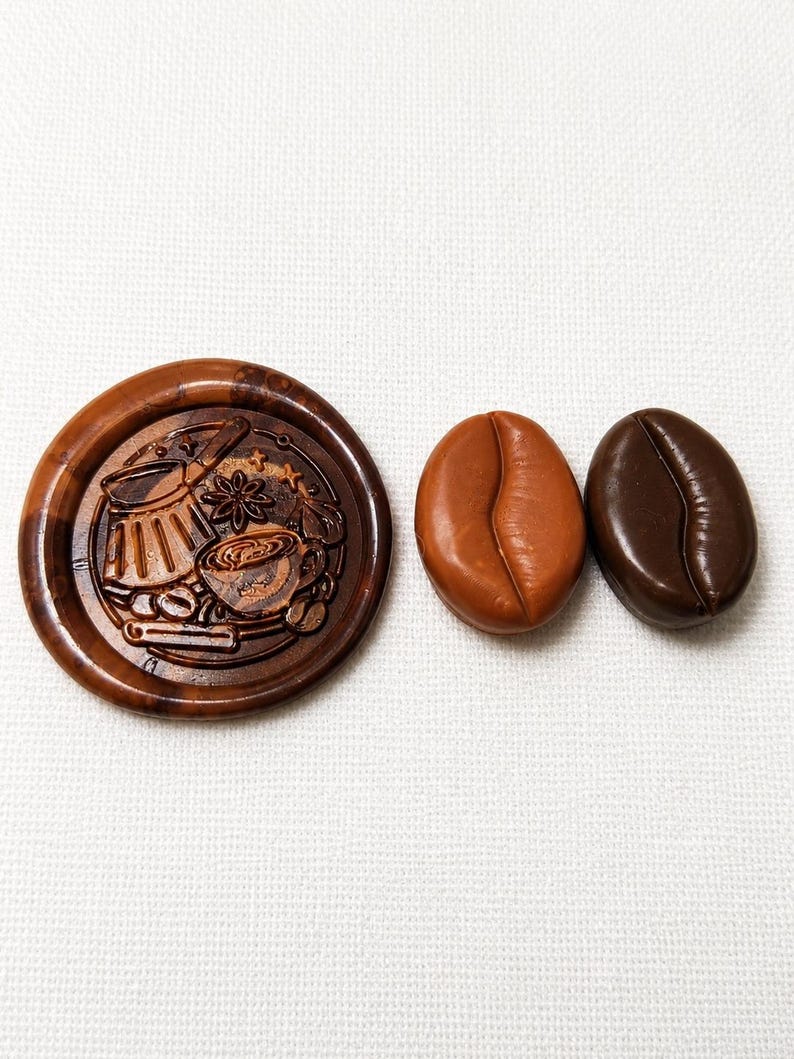May include: A circular brown wax seal stamp featuring an embossed design of a coffee pot, a cup, and spices, accompanied by two smooth, oval-shaped coffee bean replicas, one in a light tan colour and the other in a dark chocolate brown colour.