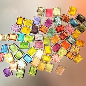 May include: A selection of rectangular watercolour paint pans in various metallic shades, such as gold, silver, pink, blue, and green. The pans are arranged randomly against a light backdrop, displaying the vibrant colours.