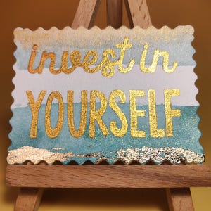 Pu&ograve; includere: Un cartello rettangolare con le parole "invest in YOURSELF" in lettere dorate. Il cartello ha uno sfondo ad acquerello con tonalit&agrave; blu e bianche ed &egrave; esposto su un piccolo cavalletto di legno.