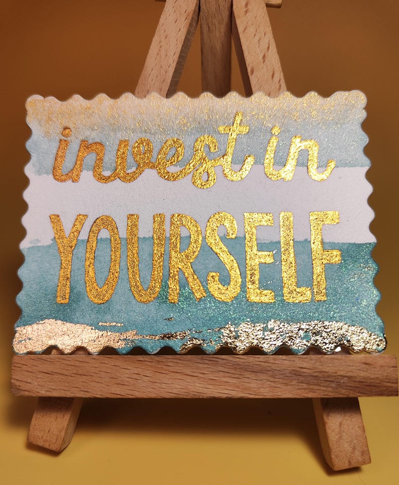 Pu&ograve; includere: Un cartello rettangolare con le parole "invest in YOURSELF" in lettere dorate. Il cartello ha uno sfondo ad acquerello con tonalit&agrave; blu e bianche ed &egrave; esposto su un piccolo cavalletto di legno.
