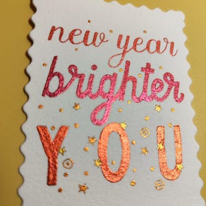 Pu&ograve; includere: Un biglietto bianco con bordo smerlato presenta le parole "new year brighter YOU" in lettere rosse e arancioni. Piccole stelle e punti dorati sono sparsi intorno al testo, creando un design festoso. Il biglietto &egrave; posto su uno sfondo giallo.