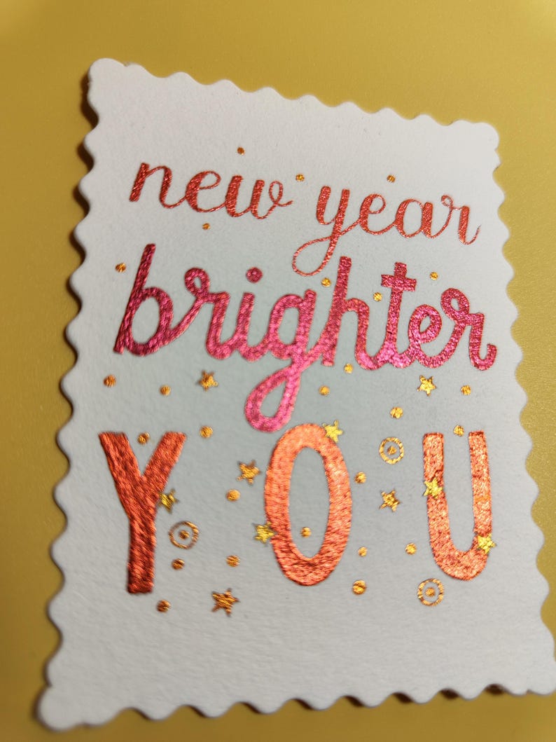 Pu&ograve; includere: Un biglietto bianco con bordo smerlato presenta le parole "new year brighter YOU" in lettere rosse e arancioni. Piccole stelle e punti dorati sono sparsi intorno al testo, creando un design festoso. Il biglietto &egrave; posto su uno sfondo giallo.