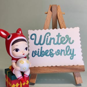 Pu&ograve; includere: Una statuina di bambola con cappuccio rosso a forma di coniglio e completo, che tiene in mano una palla bianca con disegni floreali gialli. La bambola &egrave; seduta su una scatola regalo rossa, verde e gialla. Un piccolo cavalletto di legno mostra il testo "Winter vibes only."