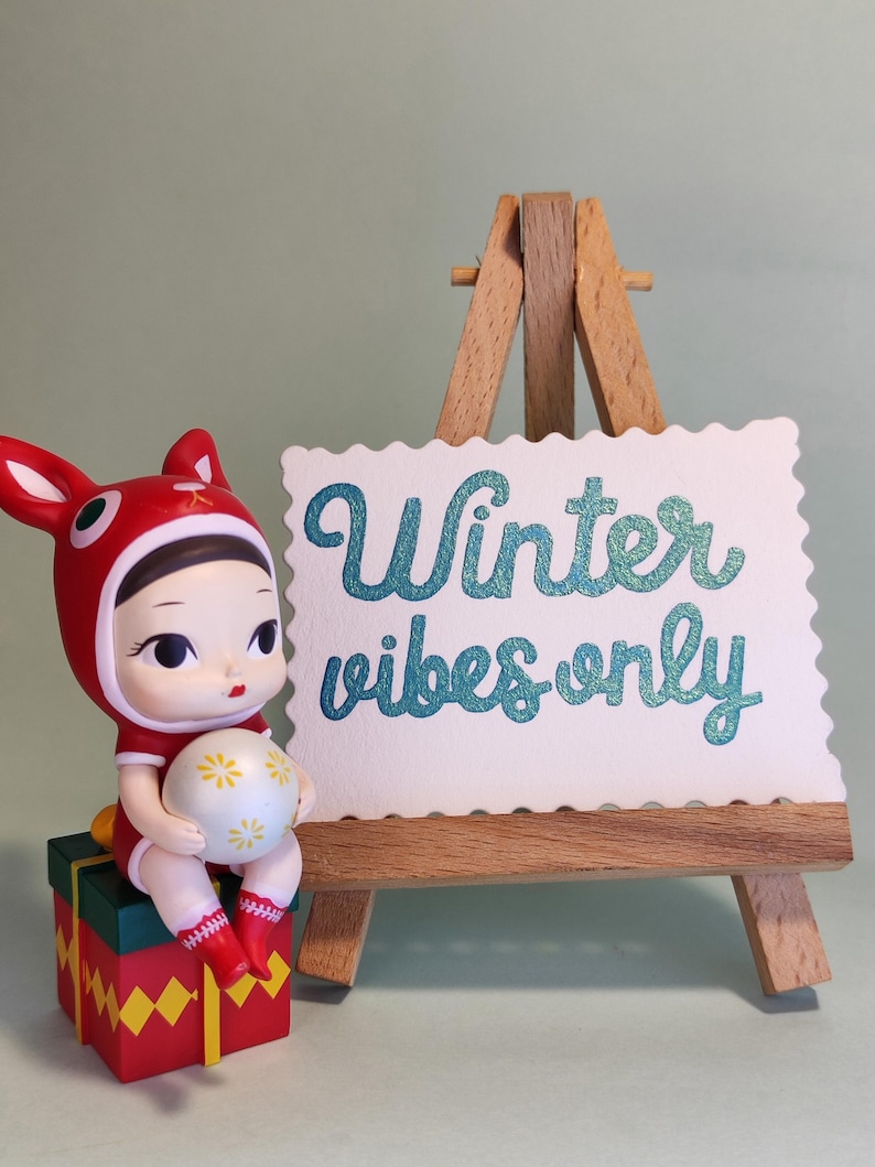 Pu&ograve; includere: Una statuina di bambola con cappuccio rosso a forma di coniglio e completo, che tiene in mano una palla bianca con disegni floreali gialli. La bambola &egrave; seduta su una scatola regalo rossa, verde e gialla. Un piccolo cavalletto di legno mostra il testo "Winter vibes only."