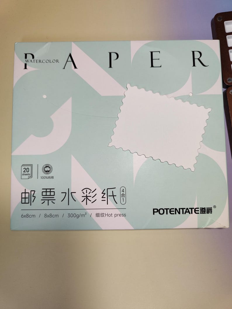 Pu&ograve; includere: Una scatola di carta per acquerello con la parola "PAPER" a lettere grandi. La scatola &egrave; verde chiaro con un ritaglio bianco a forma di francobollo. Il testo sulla scatola include "WATERCOLOR", "20 fogli" e misure di 6x8cm e 8x8cm.