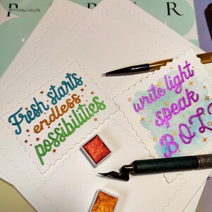 Pu&ograve; includere: Materiali per acquerello su carta bianca. L'immagine presenta due tavolozze di acquerelli, una penna nera e una penna calligrafica. La carta presenta le frasi "Fresh starts endless possibilities" e "write light speak B O L" in lettere colorate.