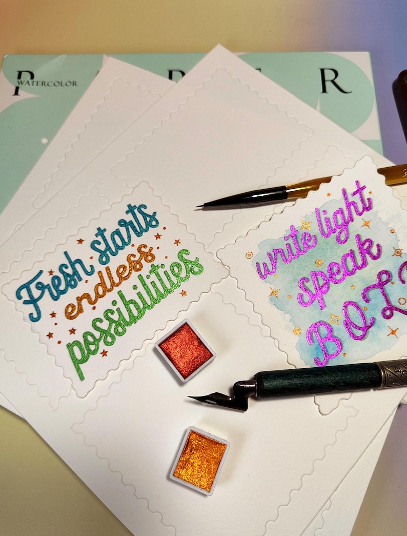 Pu&ograve; includere: Materiali per acquerello su carta bianca. L'immagine presenta due tavolozze di acquerelli, una penna nera e una penna calligrafica. La carta presenta le frasi "Fresh starts endless possibilities" e "write light speak B O L" in lettere colorate.