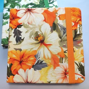 Könnte beinhalten: Zwei Notizbücher mit floralen Mustern. Das vordere Notizbuch zeigt ein lebhaftes orange-weißes Hibiskusblütenmuster mit einem orangefarbenen Gummiband. Das zweite Notizbuch hat ein grün-weißes Blumenmuster und ein grünes Gummiband.