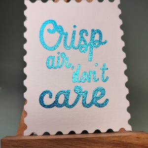 Pu&ograve; includere: Una carta bianca con bordo smerlato, con la scritta "Crisp air, don't care" in lettere turchesi scintillanti. La carta &egrave; esposta su un piccolo cavalletto di legno.