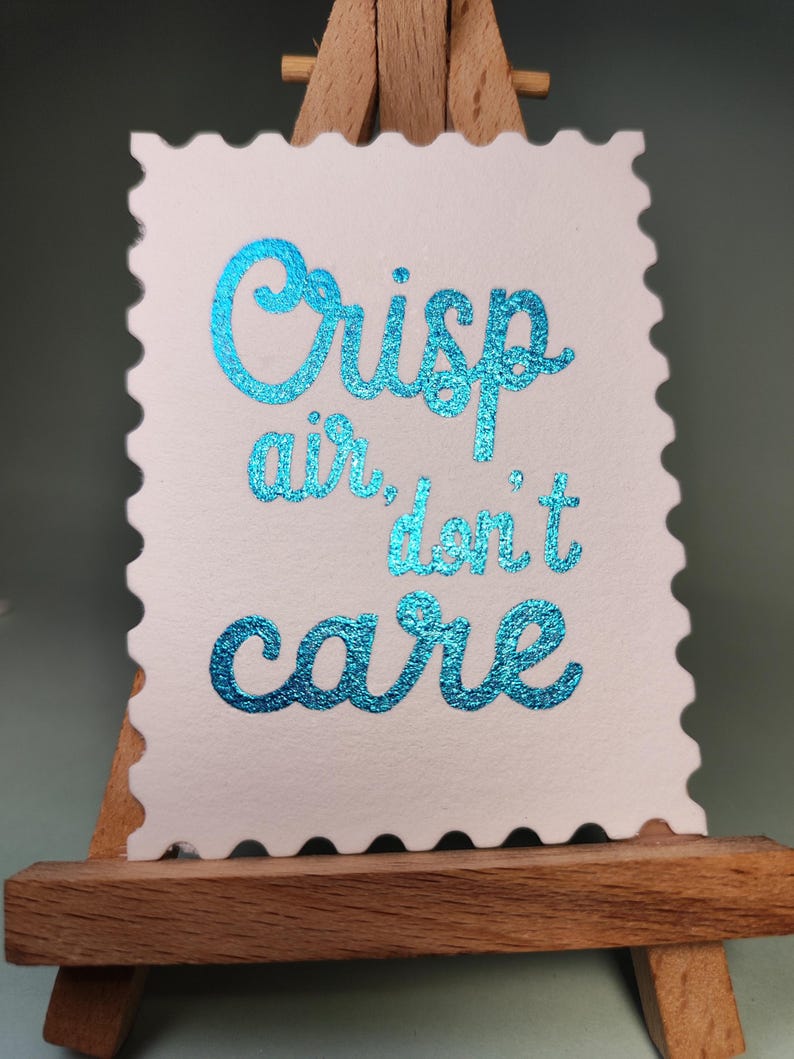 Pu&ograve; includere: Una carta bianca con bordo smerlato, con la scritta "Crisp air, don't care" in lettere turchesi scintillanti. La carta &egrave; esposta su un piccolo cavalletto di legno.
