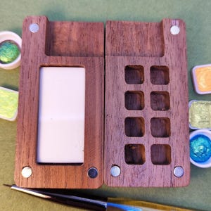 May include: A wooden watercolor paint palette, open to reveal a white mixing area and paint wells. The palette is made of dark wood and has magnetic closures. Several small paint pans with colorful paints and a fine-tipped brush are also visible.