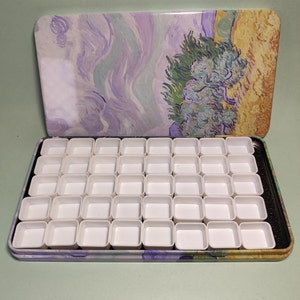 Op de afbeelding: Een open, rechthoekige aquarelverfpaletdoos met een deksel met een schilderij-achtig ontwerp in tinten paars, geel en groen. De binnenkant bevat 48 witte, vierkante verfputjes voor aquarelverf.