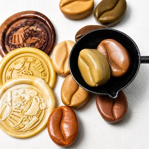 May include: A collection of smooth, coffee bean-shaped objects in various shades of brown, tan, and beige, alongside three circular wax seals featuring embossed coffee-themed designs, with two bean shapes resting inside a small black metal ladle.