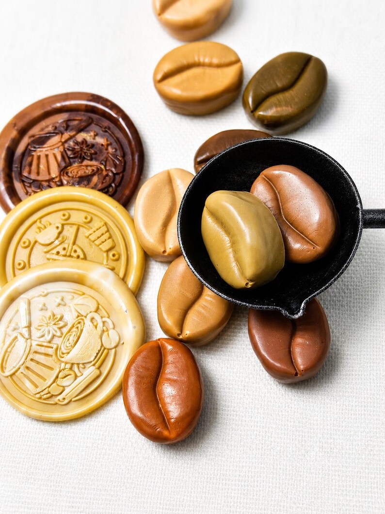 May include: A collection of smooth, coffee bean-shaped objects in various shades of brown, tan, and beige, alongside three circular wax seals featuring embossed coffee-themed designs, with two bean shapes resting inside a small black metal ladle.