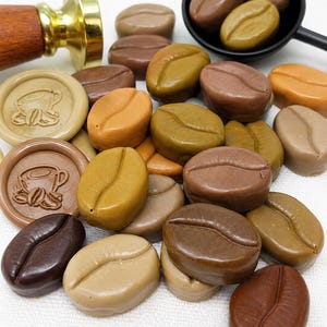 May include: A collection of wax seal beads shaped like coffee beans in various shades of brown, tan, and olive green. Two round wax seals feature a coffee cup design. A brass stamp with a wooden handle and a black scoop are also visible.