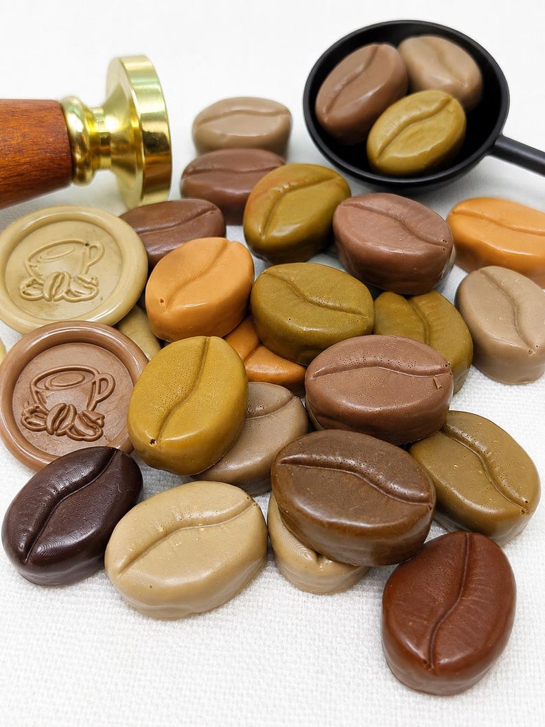 May include: A collection of wax seal beads shaped like coffee beans in various shades of brown, tan, and olive green. Two round wax seals feature a coffee cup design. A brass stamp with a wooden handle and a black scoop are also visible.