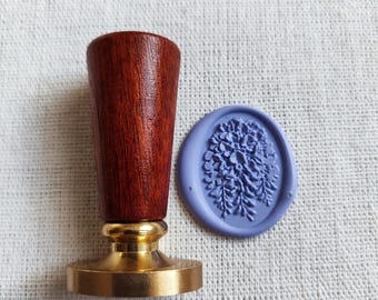 Può includere: Un timbro per ceralacca con manico in legno marrone scuro e base in metallo color oro lucido. Accanto al timbro si trova un'impronta di cera ovale color viola chiaro che mostra un dettagliato motivo floreale in rilievo.