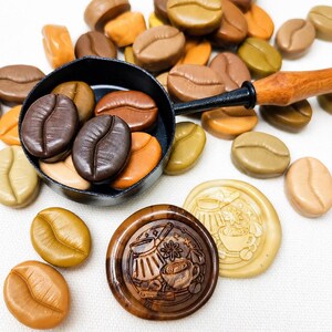 May include: A black metal melting spoon with a wooden handle containing dark brown coffee bean-shaped wax seals. Surrounding the spoon are various coffee bean-shaped wax seals in shades of tan, brown, and olive, plus two round wax seals.