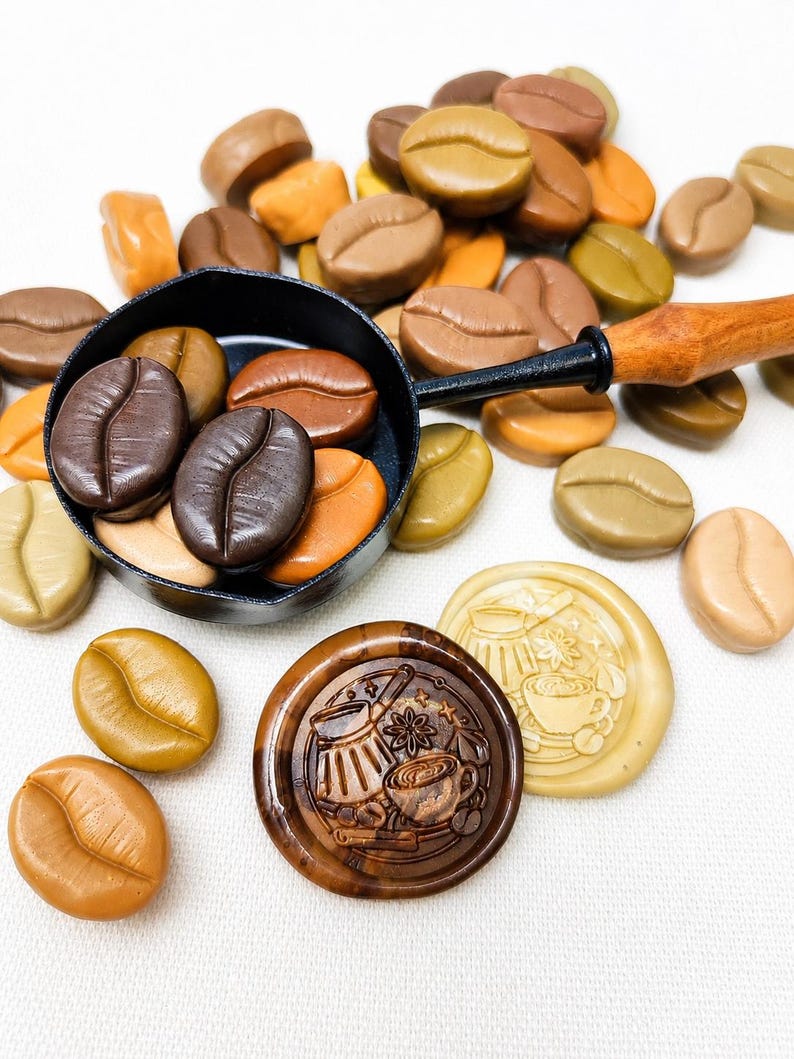 May include: A black metal melting spoon with a wooden handle containing dark brown coffee bean-shaped wax seals. Surrounding the spoon are various coffee bean-shaped wax seals in shades of tan, brown, and olive, plus two round wax seals.