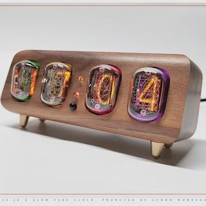 May include: A wooden digital clock with four glowing tubes displaying numbers. The clock has a rectangular walnut-coloured body with rounded edges, supported by three small, gold-coloured legs. The tubes glow in various colours, including green, orange, and purple.