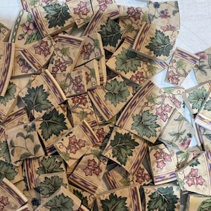 May include: A collection of broken ceramic tile pieces featuring a cream background with a repeating pattern of green leaves, small pink flowers, and thin purple and gold border stripes. The shards vary in shape and size, displaying fragments of the floral design across their surfaces.