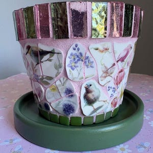 May include: Pink mosaic flower pot with a green saucer. The pot features a mosaic rim and is decorated with bird and floral designs. The saucer is a solid green color, providing a contrasting base for the pot.