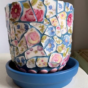 May include: A blue ceramic flower pot with a mosaic design. The mosaic is made of broken pieces of china with floral patterns in pink, red, and blue. The pot sits on a matching blue saucer.