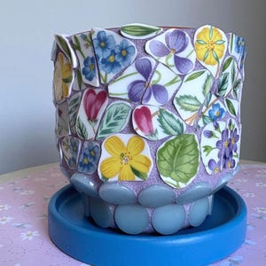 May include: A mosaic-style flower pot, decorated with floral ceramic pieces in blue, yellow, purple, and pink. The pot sits on a blue saucer, with a base of light blue stones. A unique, decorative item. The pot is approximately 15 cm in height.