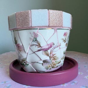 May include: A mosaic-style flower pot with a pink rim and matching saucer. The pot features a floral and bird design, with a mosaic pattern of white and pink tiles around the top.