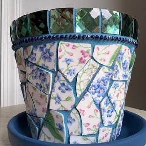 May include: A mosaic flower pot featuring white ceramic shards with blue and pink floral patterns set in blue grout. The rim is decorated with dark green reflective square tiles and a row of small dark blue beads, resting on a solid blue saucer.