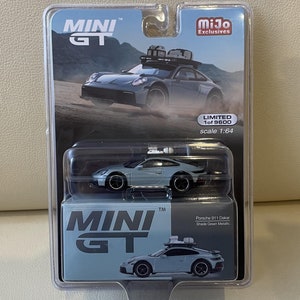 May include: A light blue Porsche 911 Dakar model car in a plastic display case. The car has a roof rack with luggage on top. The case has a Mini GT logo and the text "Limited 1 of 9600" and "scale 1:64".
