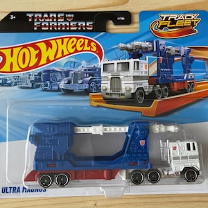 Pode incluir: Um camião de brincar Hot Wheels Transformers Ultra Magnus na embalagem original. O veículo é branco e azul com detalhes em vermelho, apresentando um design de reboque de plataforma. O cartão exibe os logótipos Transformers e Track Fleet.