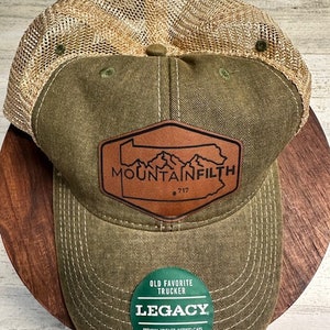 Peut inclure: Un chapeau de camionneur vert et marron avec un patch en cuir marron qui dit "MOUNTAINFILTH 717". Le chapeau a un dos en maille et un autocollant vert de la marque Legacy.