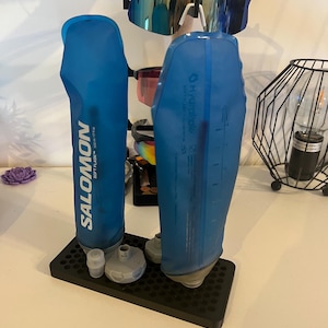 Peut inclure: Un support de séchage en plastique noir contenant deux flasques souples bleues Salomon de 500 ml, une paire de lunettes de soleil à monture blanche avec des verres réfléchissants et d'autres lunettes. Le support possède une base perforée.