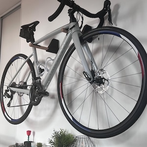 Può includere: Una bici da corsa grigio chiaro montata su una parete bianca. La bici ha manubrio nero, sella nera e ruote con dettagli rossi e blu. La bici è esposta sopra una mensola con piccole piante e una lampada decorativa.