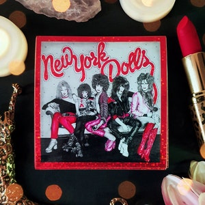 Puede incluir: Una caja cuadrada con borde rojo y la inscripción "New York Dolls" en letras rojas. La imagen muestra a cinco personas con ropa glam rock. Un labial rojo en un estuche dorado con estampado de leopardo está junto a la caja.