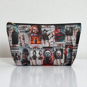 Può includere: Un beauty case nero con cerniera con un tema di film horror, che mostra un collage di personaggi e titoli di giornali. Il design include immagini di Freddy Krueger, Chucky e Michael Myers, con testo come "House of Horrors."